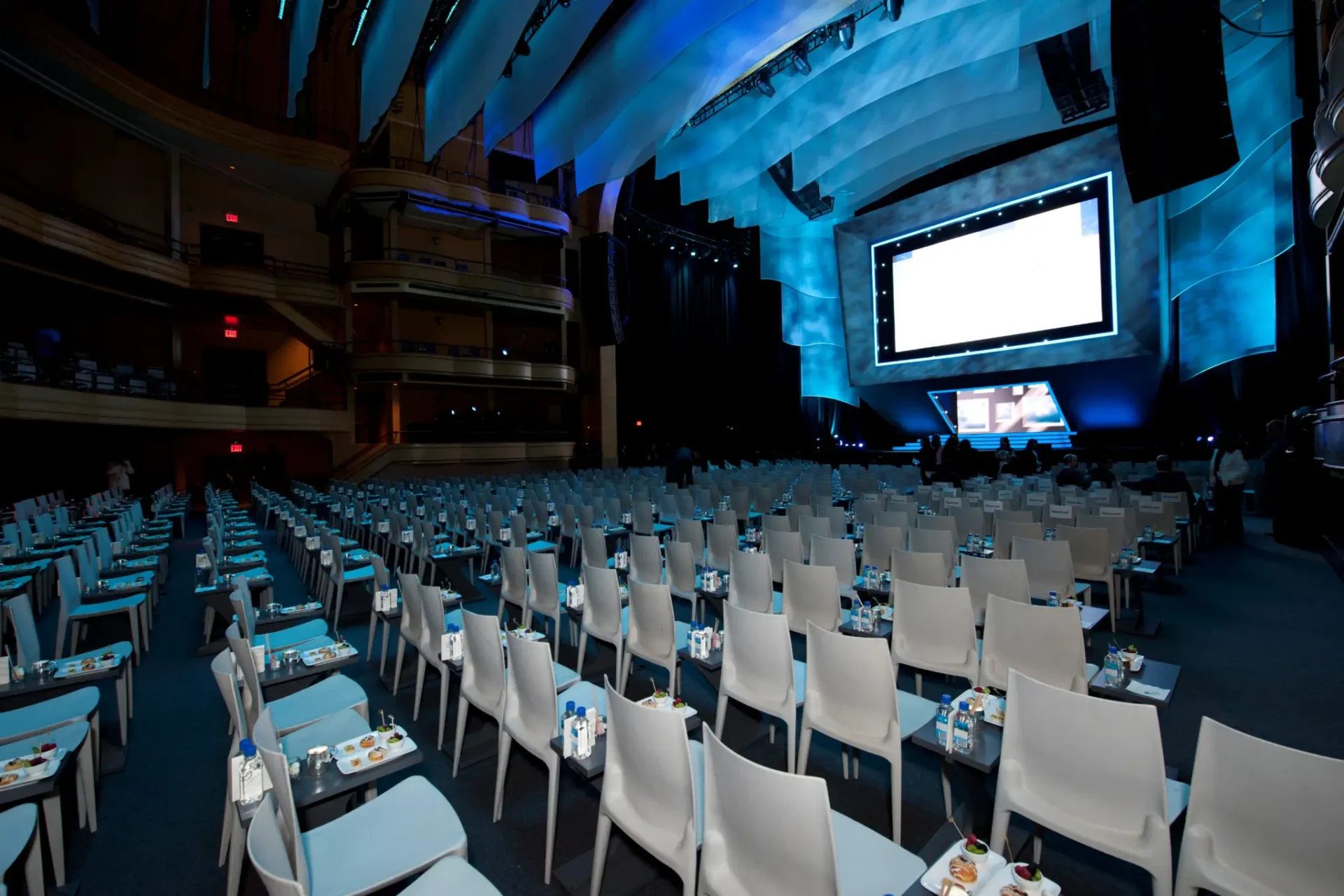 An empty conference hall with rows of chairs, tables set with water and notepads, and a large presentation screen onstage