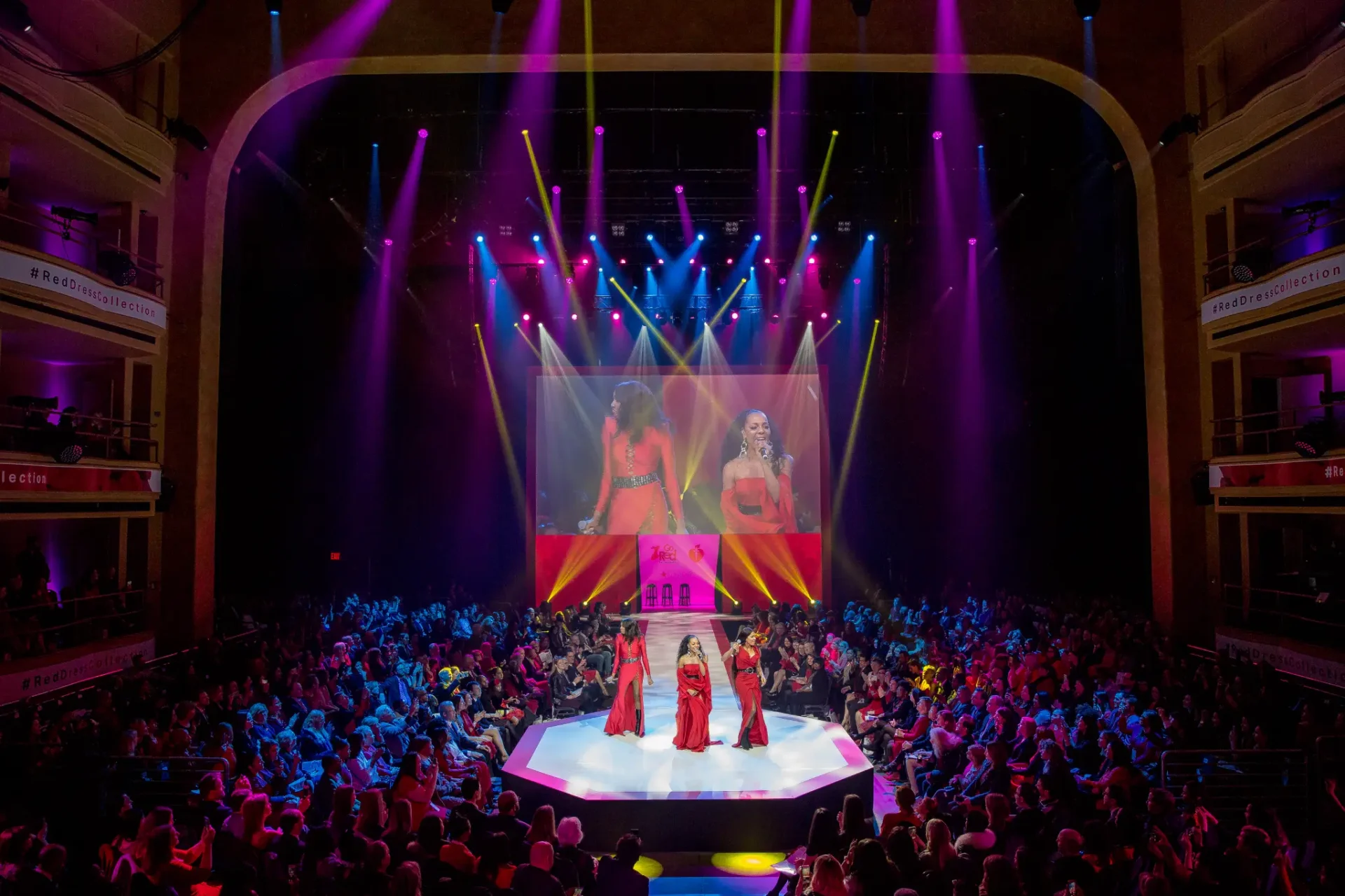 Fashion show with models on runway, audience attentively seated, and colorful stage lights